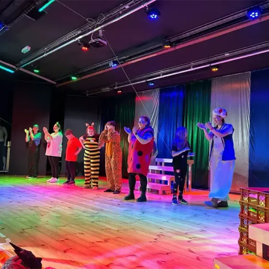 Eine Gruppe von Darstellern auf der Bühne unter buntem Licht. Einige tragen Kostüme, andere lässige Kleidung. Sie scheinen zu klatschen oder zu jubeln.