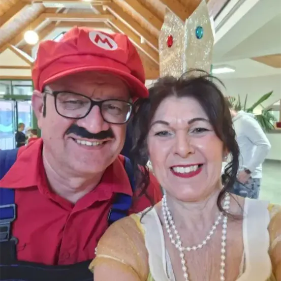 Ein Mann und eine Frau lächeln und posieren für ein Foto. Der Mann trägt einen roten Hut und einen Schnurrbart. Die Frau trägt eine Halskette. Dahinter befindet sich ein verschwommener Hintergrund einer Person.