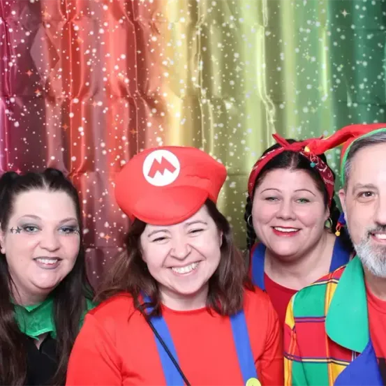 Fünf Menschen in bunten Kostümen posieren für ein Foto vor einem mehrfarbigen Hintergrund. Eine Person trägt einen Mario-Hut.