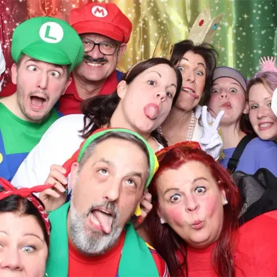 Eine Gruppe von Menschen in Kostümen posiert für ein Foto in einer farbenfrohen Umgebung. Einige tragen Mario- und Luigi-Hüte, andere rote Perücken. Sie machen lustige Gesichter und stecken die Zungen heraus.