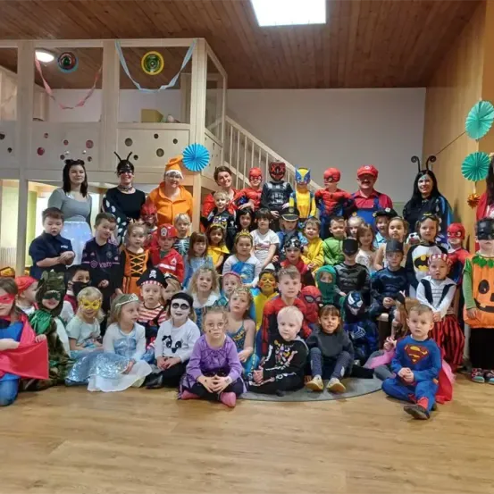 Eine Gruppe von Kindern und Erwachsenen in Kostümen posiert für ein Foto in einem Raum mit Holzboden, Dekorationen und einer Treppe.