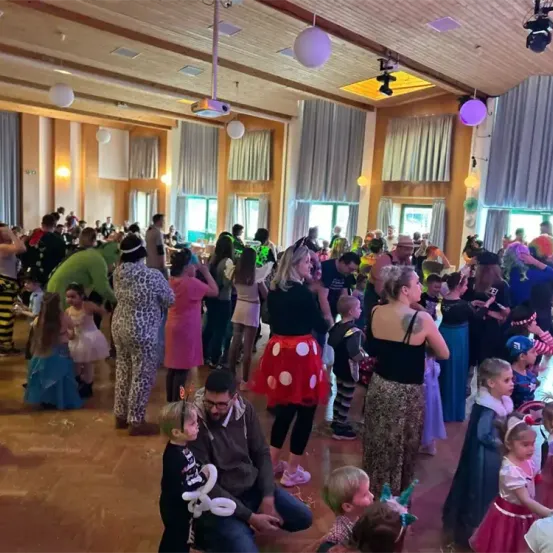 Ein überfüllter Raum mit Menschen in Kostümen, darunter Kinder und Erwachsene, die tanzen und eine festliche Veranstaltung genießen. Der Raum hat Holzböden, hängende Lichter und Fenster.