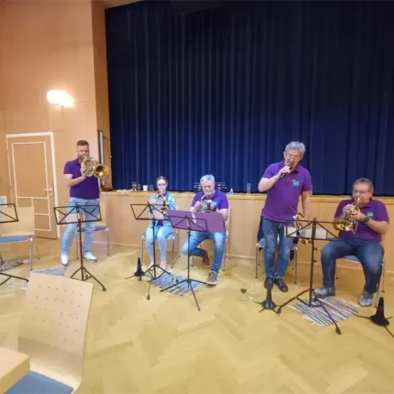 Fünf Musiker in lila Hemden spielen Trompeten in einem Saal mit blauem Bühnenvorhang. Vier sitzen auf Stühlen, einer steht.