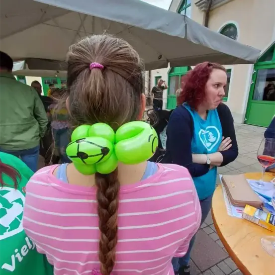 Eine Frau mit grünen Luftballon-Haargummis steht unter einem Schirm. Eine Frau mit roten Haaren und einem blauen Shirt steht neben ihr. Ein Tisch mit einem Weinglas und einem Buch ist in der Nähe.