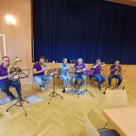 Sechs Männer in lila Hemden sitzen auf Stühlen in einem Raum und spielen Blechblasinstrumente. Sie stehen vor einer Bühne mit einem blauen Vorhang.