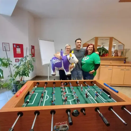 Drei Personen stehen hinter einem Tischkicker. Einer hält eine Kuh-Plüschfigur. Dahinter sind ein Whiteboard und Pflanzen zu sehen. Rechts steht ein Schrank mit einer Pflanze.