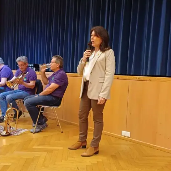 Eine Frau steht und hält ein Mikrofon. Drei Männer sitzen hinter ihr. Einer der Männer trinkt Wasser, während die anderen beiden eine Trompete halten. Sie tragen alle lila Hemden. Dahinter befindet sich ein blauer Vorhang.