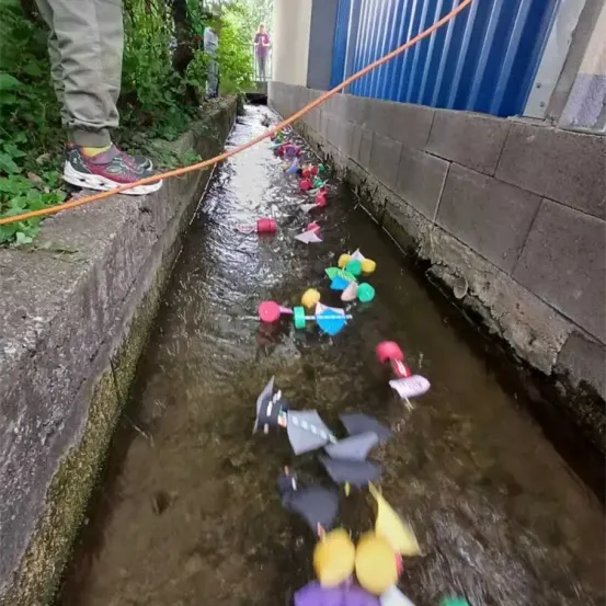Eine Person steht in der Nähe eines Betonrandes und beobachtet bunte schwimmende Objekte in einem Bach. Verschiedene Spielzeuge und Gegenstände schwimmen im Wasser.