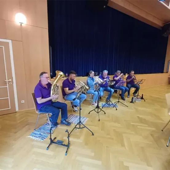 Eine Gruppe von Musikern in lila Hemden spielt Blechblasinstrumente in einem Raum mit Holzböden und einem blauen Vorhang im Hintergrund.