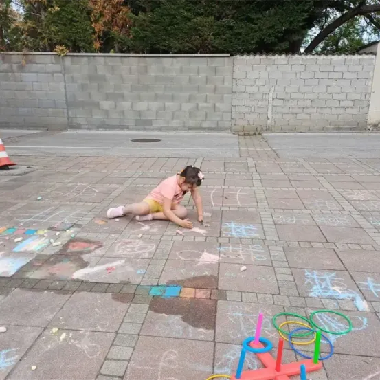 Ein kleines Mädchen malt mit Kreide auf dem Pflaster in der Nähe einer Mauer, mit verschiedenen farbigen Kreiden und Spielzeug verstreut um sie herum.