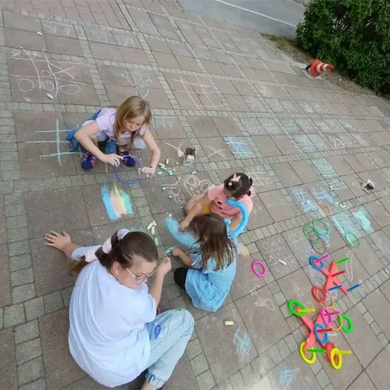 Vier junge Mädchen spielen Hopscotch und zeichnen mit Kreide auf einem Ziegelboden. Eine hält eine Hula-Hoop. Zeichnungen und Spielzeug liegen auf dem Boden verstreut.