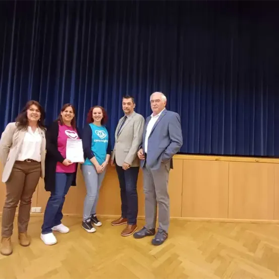 Fünf Menschen stehen lächelnd zusammen auf einer Bühne. Dahinter ein blauer Vorhang und Holzpaneele. Eine Frau hält ein Zertifikat. Eine andere Frau trägt ein rosa T-Shirt mit Herzdesign.