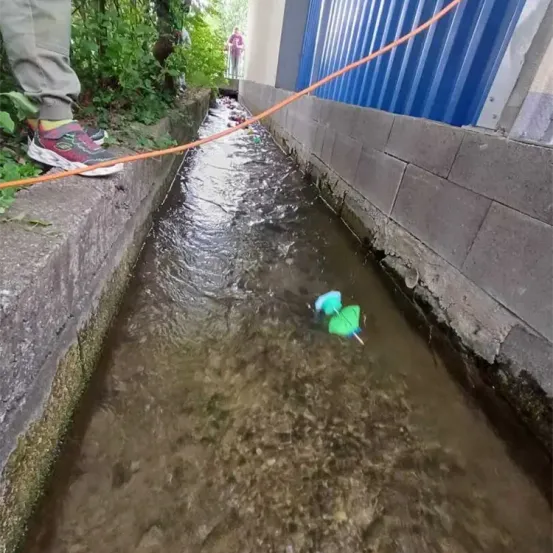 Eine Person in Turnschuhen steht neben einem fließenden Bach, ein orangefarbener Schlauch erstreckt sich darüber. Ein grüner Dart schwimmt im Wasser, während sich jemand im Hintergrund bückt.