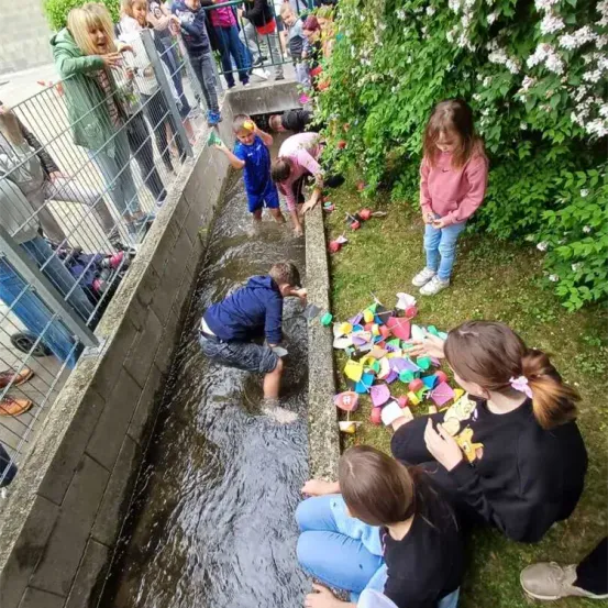 Mehrere Kinder spielen in einem Kanal und setzen schwimmende Objekte ins Wasser. Einige Kinder stehen im Wasser, während andere auf dem Gras sitzen. Hinter ihnen beobachten mehrere Menschen aus einem eingezäunten Bereich.