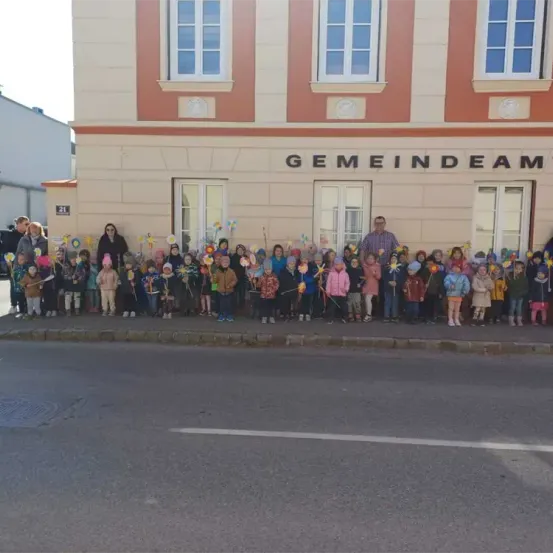 Eine Gruppe von Kindern und Erwachsenen steht vor einem Gebäude mit dem Namen 'GEMEINDEAM' und hält bunte Objekte in der Hand. Das Gebäude hat mehrere Fenster und befindet sich in der Nähe einer Straße.