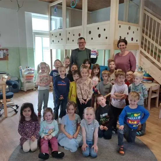 Eine Gruppe von Kindern und zwei Erwachsenen sind in einem Klassenzimmer und lächeln für ein Foto. Die Erwachsenen halten ein Buch. Der Raum hat Holzböden, Schränke und eine Treppe.
