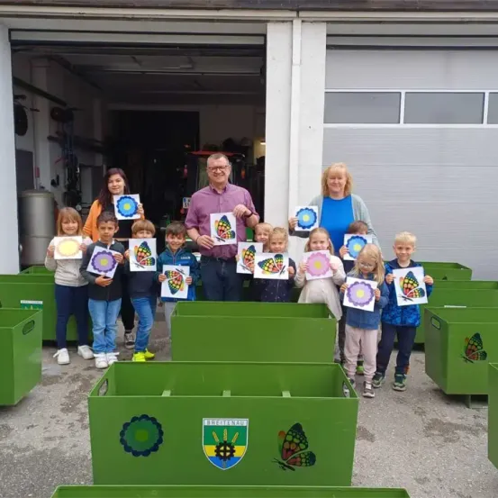 Eine Gruppe von Kindern und Erwachsenen hält bunte Schmetterlingszeichnungen vor grünen Aufbewahrungsboxen und steht im Freien vor einer Garage.