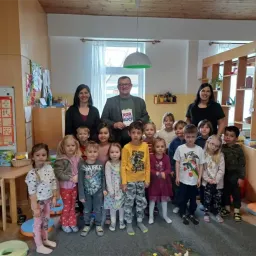 Eine Gruppe von Kindern, Erwachsenen und einem Mann, der ein Buch mit dem Wort 'vor' in der Hand hält, steht in einem Raum. Dahinter befindet sich ein Regal mit Gegenständen. An der Wand sind Papiere befestigt. Es gibt einen Tisch mit einem Korb oben.