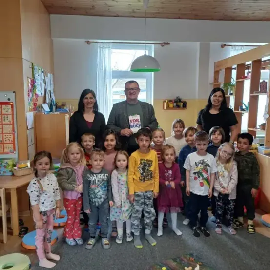 Eine Gruppe von Kindern, Erwachsenen und einem Mann, der ein Buch mit dem Wort 'vor' in der Hand hält, steht in einem Raum. Dahinter befindet sich ein Regal mit Gegenständen. An der Wand sind Papiere befestigt. Es gibt einen Tisch mit einem Korb oben.