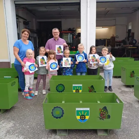 Eine Gruppe von Kindern und Erwachsenen steht vor grünen Kisten und hält Bilder von Schmetterlingen und Blumen hoch. Die Umgebung scheint eine Werkstatt oder eine Garage zu sein.