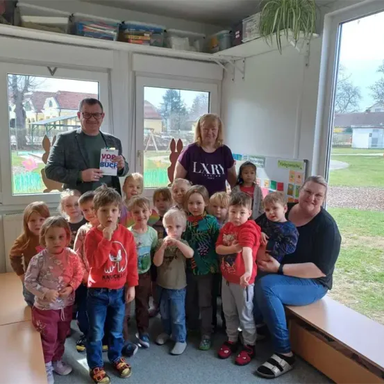 Eine Gruppe junger Kinder und Erwachsener posiert in einem Raum für ein Foto und lächelt. Ein erwachsener Mann hält ein Buch und eine Frau trägt ein lila Shirt. Der Raum hat Glasfenster und Regale mit verschiedenen Gegenständen. Draußen gibt es einen Rasenbereich mit Bäumen und einem Gebäude.