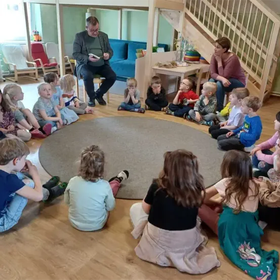 Ein Mann liest ein Buch vor einer Gruppe von Kindern, die in einem Kreis sitzen. Die Kinder sehen interessiert und engagiert aus.
