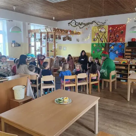 Ein Klassenzimmer mit Kindern, die um einen Tisch herumsitzen, ein Mann in einem grünen Kostüm steht vor ihnen und farbenfrohe Gemälde schmücken die Wände.