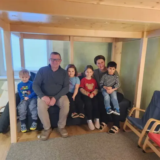 Eine Familie sitzt auf einem Sofa in einem Raum mit Holzrahmen. Zwei Frauen, ein Mann und vier Kinder lächeln. Sie tragen Freizeitkleidung. Der Raum hat einen Teppich und ein Fenster auf der linken Seite.