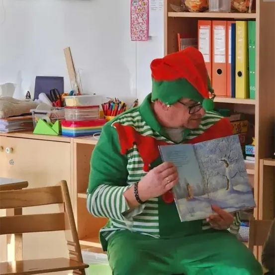 Eine als Elf verkleidete Person sitzt in einem Klassenzimmer und liest ein Buch. Der Raum ist organisiert mit Regalen gefüllt mit Ordnern und Zubehör.
