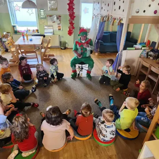 Ein Clown sitzt auf einem Hocker in einem Raum, umgeben von Kindern auf Kissen. Der Raum hat Holzböden, einen Tisch und Stühle.
