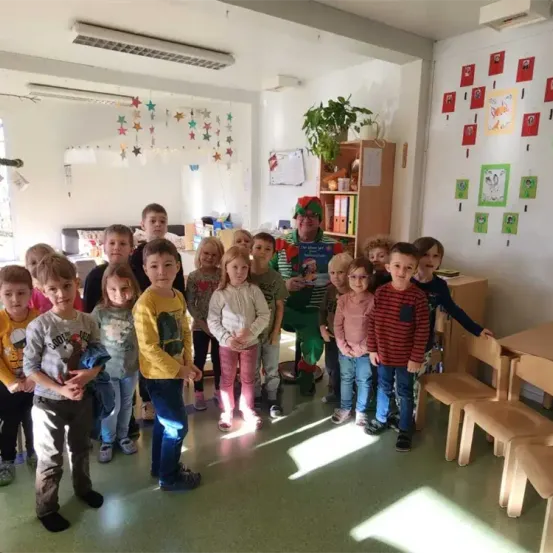 Ein Klassenzimmer mit mehreren Kindern, die zusammenstehen, ein Mann in festlicher Kleidung mit einem Buch, Topfpflanzen und dekorierten Wänden.
