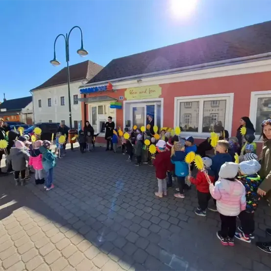 Eine Gruppe von Kindern mit Sonnenblumen steht vor einem Gebäude mit einem orangefarbenen Schild, auf dem 'Klein und Fein' steht. Eine Person steht in der Nähe des Eingangs.