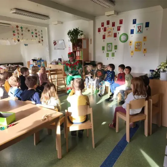 Ein Klassenzimmer mit Kindern, die um einen als Weihnachtsmann verkleideten Mann herumsitzen, der ein Buch liest, und farbenfrohen Dekorationen an den Wänden.