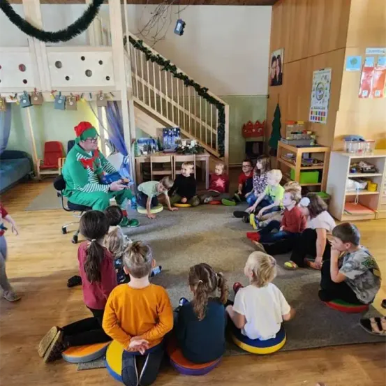 Kinder in einem Klassenzimmer lauschen einem Mann, der als Weihnachtsmann verkleidet ist. Der Raum ist weihnachtlich geschmückt.