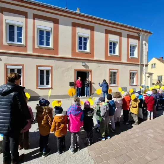 Eine Gruppe von Kindern, die bunte Mäntel und Mützen tragen, steht vor einem Gebäude. Sie halten Sonnenblumen. Zwei Erwachsene stehen in der Tür des Gebäudes. Das Gebäude hat mehrere Fenster und einen Schornstein.