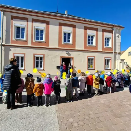 Eine Gruppe von Kindern mit Sonnenblumen steht vor einem Gebäude mit mehreren Fenstern und einer offenen Tür.