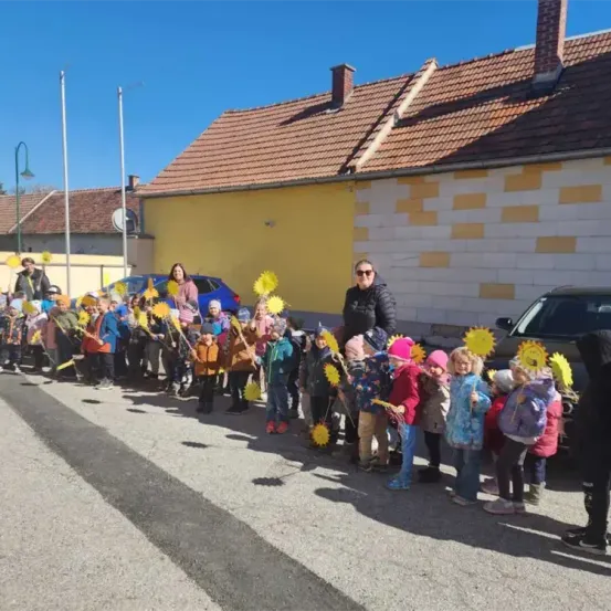 Eine Gruppe von Kindern hält Sonnenblumen vor einem Gebäude. Ein Erwachsener steht neben ihnen. Vor dem Gebäude sind Autos geparkt.