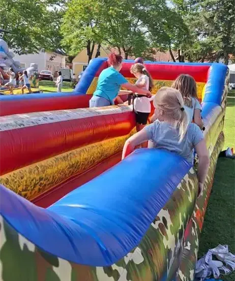 Eine Gruppe von Kindern klettert auf einen mehrfarbigen aufblasbaren Rutschvergnügen. Eine Erwachsene unterstützt ein Mädchen in der Nähe der Spitze.