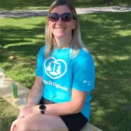 Eine Frau mit Sonnenbrille und blauem T-Shirt sitzt lächelnd auf einer Parkbank.