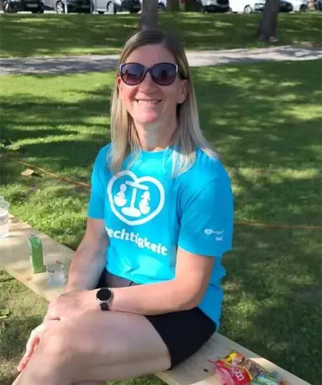 Eine Frau mit Sonnenbrille und blauem T-Shirt sitzt lächelnd auf einer Parkbank.