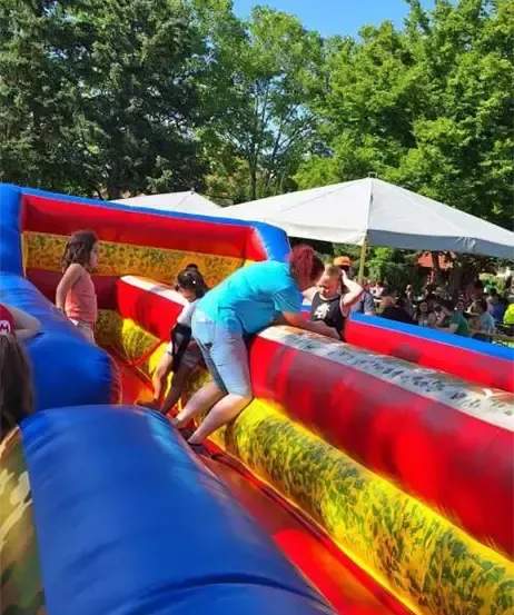 Ein Outdoor-Event mit einem großen aufblasbaren Rutschen. Kinder und Erwachsene spielen darauf. Ein Erwachsener hilft den Kindern beim Klettern auf den Rutsch. Leute sitzen unter einem Zelt. Bäume sind im Hintergrund.