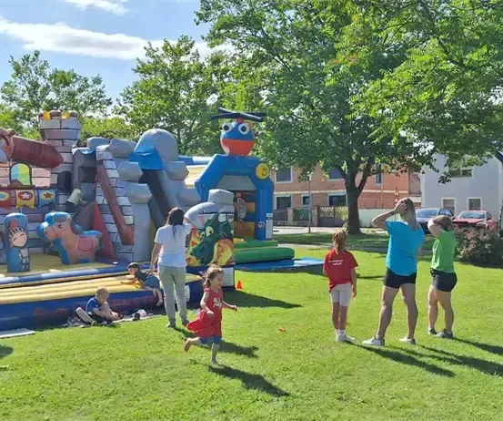Eine Gruppe von Kindern und Erwachsenen spielt in einem Außenbereich mit Hüpfburgen und einer Rutsche. Einige sitzen und andere stehen. Bäume und Häuser sind im Hintergrund.