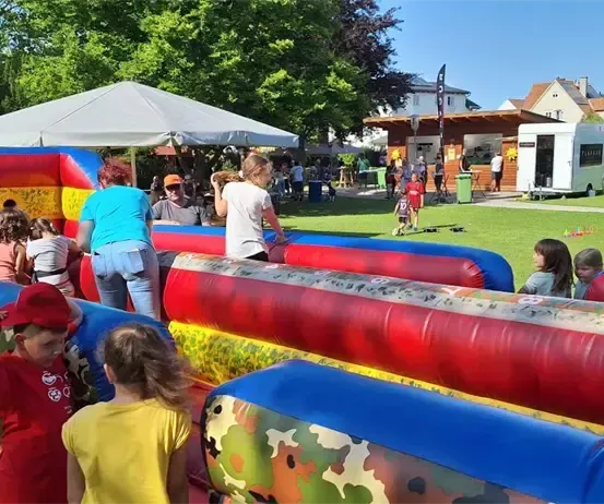 Kinder spielen auf einem aufblasbaren Hindernisparcours im Freien, mit Erwachsenen und anderen Kindern in der Nähe. Im Hintergrund befinden sich Zelte, ein Gebäude und ein geparkter Van.