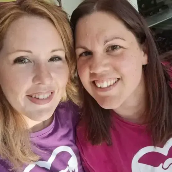 Zwei Frauen, beide mit herzförmigen Grafik-T-Shirts, lächeln und posieren für ein Foto.