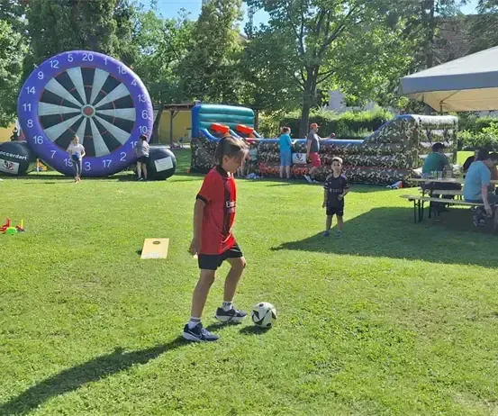 Ein Junge tritt einen Fußball auf einem grünen Feld mit aufblasbaren Spielen und einem Zelt im Hintergrund.