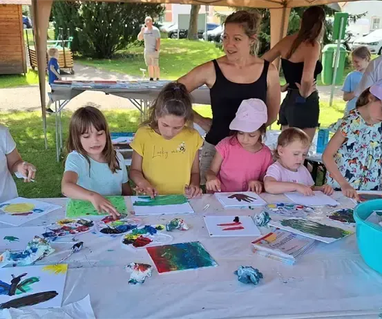 Eine Gruppe von Kindern malt mit einer Erwachsenen. Sie sitzen um einen Tisch herum, der mit Papieren und Farbe bedeckt ist. Der Tisch hat Pinsel, Malpaletten und eine Schüssel mit Papieren. Die Erwachsene trägt ein ärmelloses Oberteil und lächelt. Die Kinder tragen Hüte. Im Hintergrund befindet sich eine Rasenfläche mit einem Zelt und einem Tisch mit Büchern.