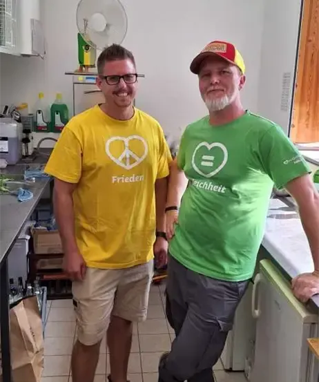 Zwei Männer posieren für ein Foto in einer Küche. Einer trägt ein gelbes T-Shirt mit dem Wort Frieden, der andere ein grünes T-Shirt mit dem Wort Gleichheit.