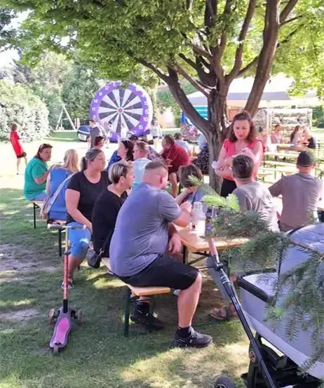 Eine Gruppe von Menschen sitzt um einen Tisch unter einem Baum. In der Nähe befinden sich ein Dartboard, ein Kinderwagen und eine Person auf einem Roller.