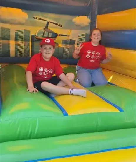 Zwei Kinder in roten T-Shirts sitzen in einem gelben und grünen aufblasbaren Hüpfhaus. Sie posieren für ein Foto, eines davon zeigt den Friedenszeichen. Der Hintergrund zeigt eine Wandmalerei mit einem Hubschrauber und einer Stadtlandschaft.