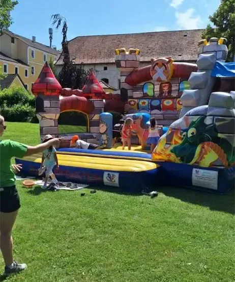 Eine Gruppe von Kindern spielt auf einem aufblasbaren Schloss auf einer Rasenfläche. Eine Frau zeigt auf sie.